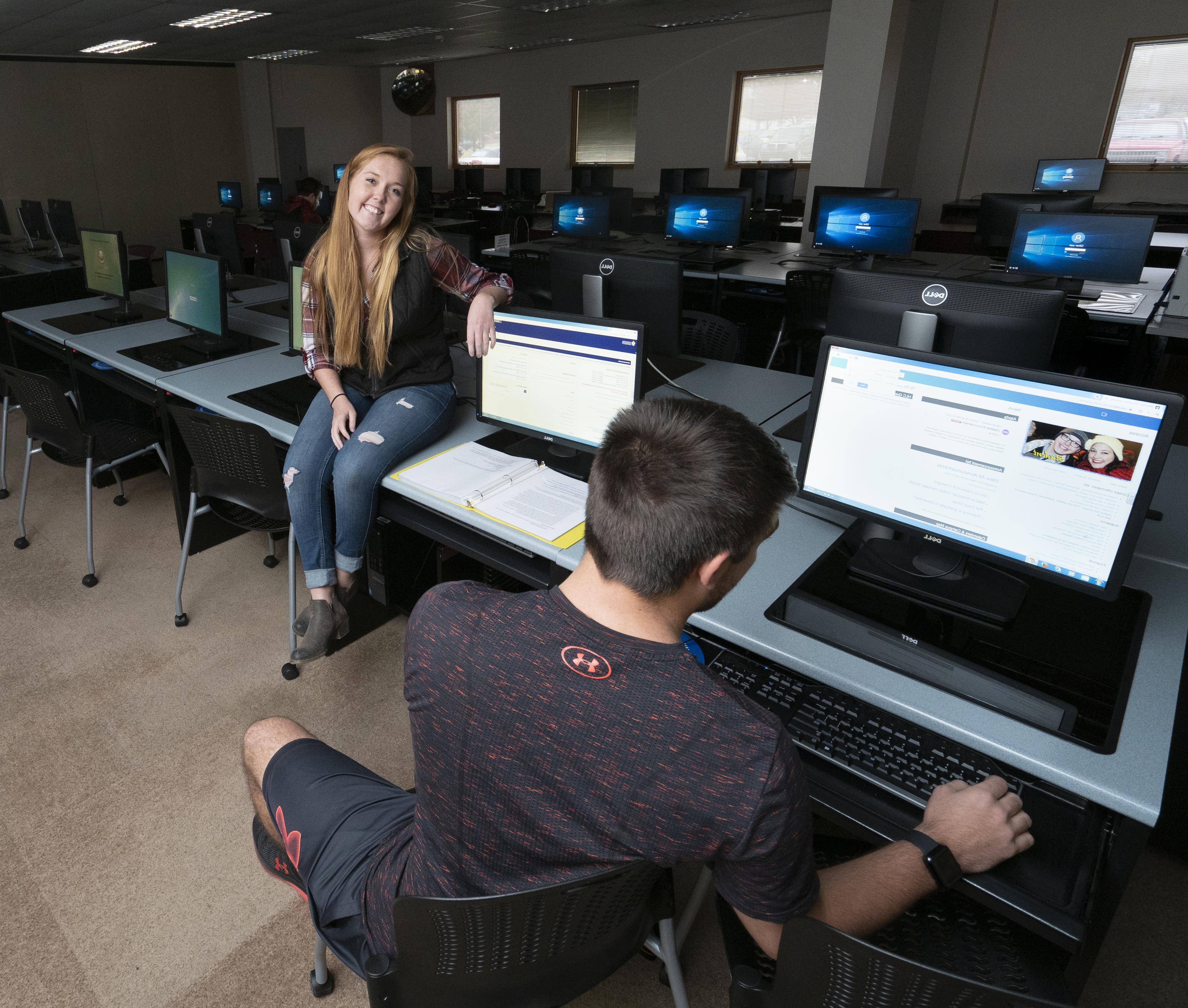 A female student sitting on a table in the computer lab while another student works